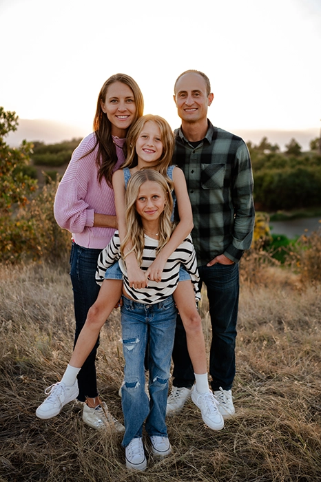 Nick Caras smiling outside with his wife and two daughters. One of his daughters is giving the other a piggyback ride.